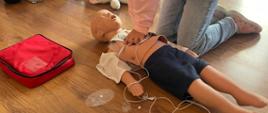 Dziewczynka wykonuje masaż serca na manekinie poprzez uciskanie na klatkę piersiową splecionymi dłońmi, inne dzieci przyglądają się tej czynności