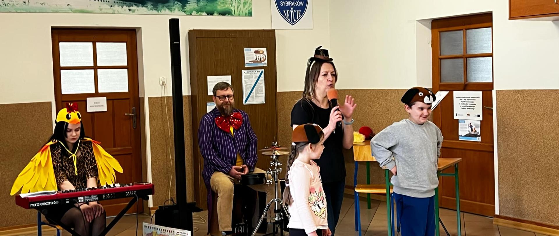 Pani ubrana w czarną koszulkę, czarne spodnie i czerwone buty trzyma w dłoni mikrofon przez który przemawia. Obok niej stoi dwoje uczniów - dziewczynka i chłopiec. Oboje mają na głowach czepeczki z maskami piesków. Za nimi siedzi Pani przebrana za kaczkę. Przed Panią stoi keyboard. Obok niej siedzi Pan przebrany za Pana Kleksa. Przed Panem stoi perkusja.