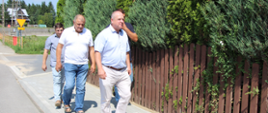 Zdjęcie ukazuje czterech mężczyzn idących po nowo wyłożonym chodniku bezpośrednio obok brązowego, drewnianego płotu i wysokich tui.