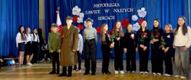 Chłopiec w wojskowym płaszczu i czapce, obok chłopiec w mundurze harcerskim. Za nimi dziewczynki stoją w rzędzie