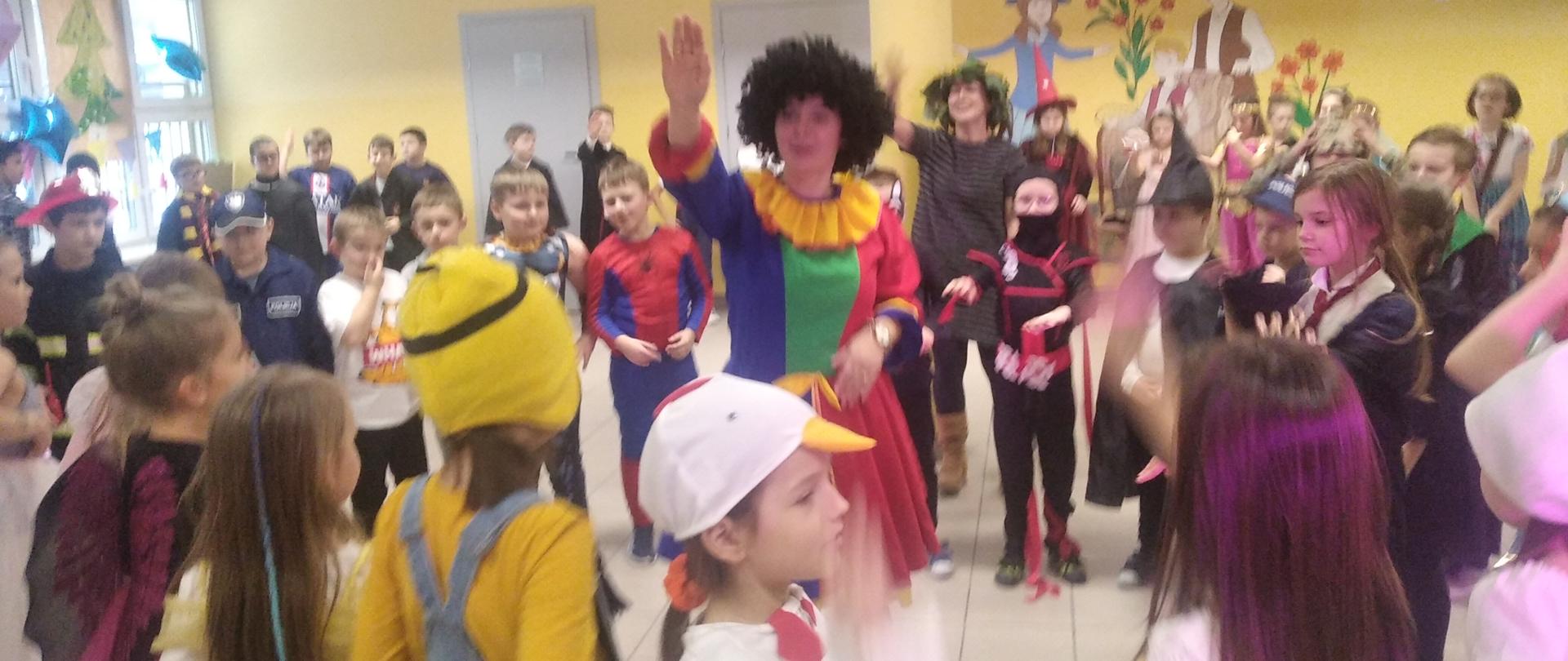 Dorosła osoba przebrana w kolorowy strój klauna pokazuje ruchy rąk do tańca macarena. Uczestnicy balu naśladują prowadzącą. 
