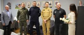 Oficjalne spotkanie w gabinecie. W rzędzie pod ścianą stoi czterech mężczyzn w różnych mundurach: wojskowym, policyjnym, strażackim oraz służby więziennej. Po lewej stronie stoi mężczyzna w szarej marynarce, a po prawej kobieta w jasnej sukience, która trzyma bukiet białych róż. Wszyscy zwróceni są w stronę kobiety. Na szarej ścianie w tle widoczne są oprawione portrety. W dolnym rogu logo strony powiatzambrowski.com.