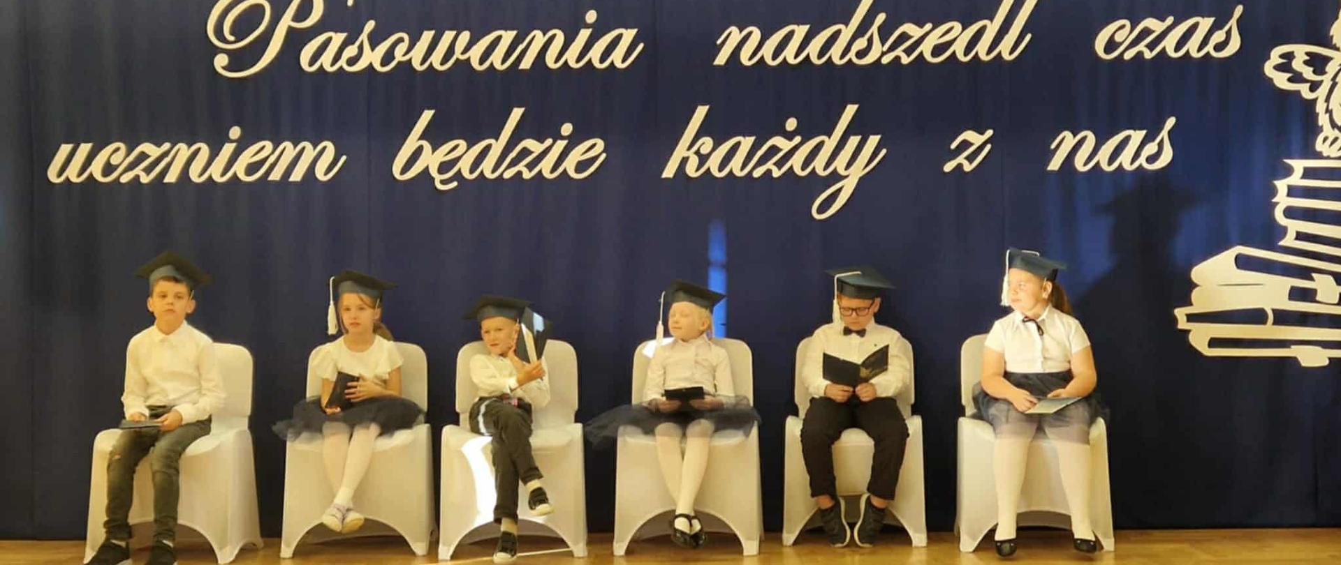 Sześcioro dzieci w galowych strojach(3 dziewczynki i 3 chłopców) siedzi na pięknie ustrojonych krzesłach na sali gimnastycznej. W tle widać dekorację - na szafirowym tle ogromnymi literami napis "Pasowania nadszedł czas, dziś uczniem będzie każdy z nas" oraz ogromna sowa nad stosem książek.