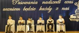 Sześcioro dzieci w galowych strojach(3 dziewczynki i 3 chłopców) siedzi na pięknie ustrojonych krzesłach na sali gimnastycznej. W tle widać dekorację - na szafirowym tle ogromnymi literami napis "Pasowania nadszedł czas, dziś uczniem będzie każdy z nas" oraz ogromna sowa nad stosem książek.