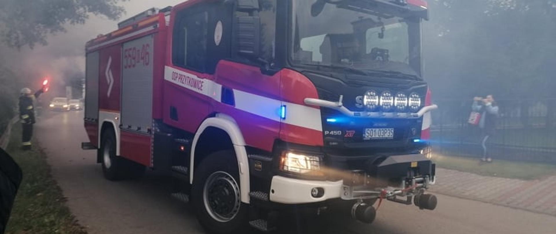 Ciężki wóz ratowniczo-gaśniczy marki Scania na asfaltowej drodze w Przytkowicach. Na poboczu strażacy z racami, powietrze za wozem zadymione od rac.