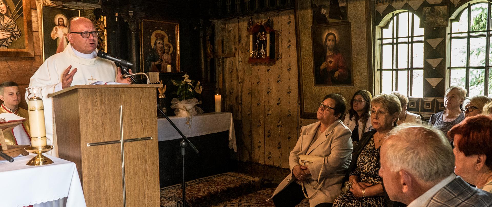 Wnętrze kaplicy z licznymi obrazami na ścianach, ksiądz w białym ornacie przemawia do siedzących wiernych.