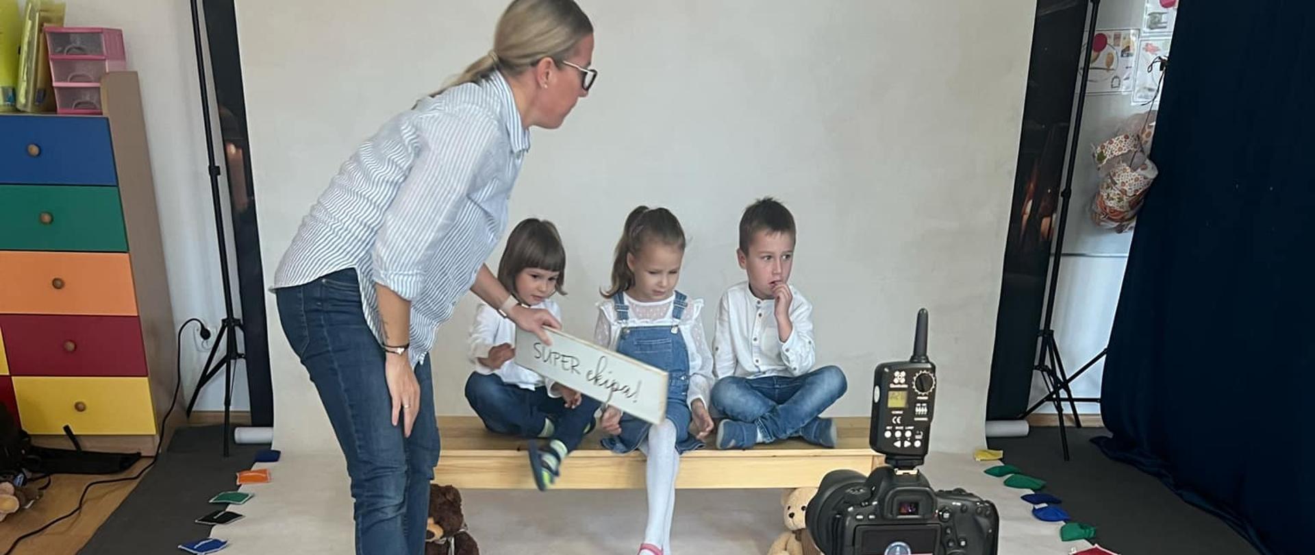 Troje dzieci siedzi na ławeczce, za nimi widoczne jest białe tło. Pani fotograf ustawia dzieci do zdjęcia, jednemu z chłopców podaje tabliczkę z napisem „Hej Przedszkole!”