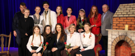 Zdjęcie przedstawia grupę osób na scenie, po premierze sztuki „Moralność Pani Dulskiej” w MOK-u. Wśród nich znajdują się dwie kobiety w centralnej części zdjęcia, które trzymają czerwone róże. Na środku znajduje się Starosta Zambrowski, Edyta Marchelska-Groszfeld. Pozostałe osoby to uczniowie, aktorzy i nauczyciele z ZS1 w Zambrowie. W tle widoczna jest scenografia z namalowanym kominkiem i zasłonami.