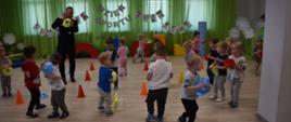 Na zdjęciu widnieją dzieci z grupy Tygryski wraz z trenerem. Prócz dzieci widać pachołki oraz inne akcesoria pomocne przy treningach. W tle widnieje napis " Dzień Sportu" oraz ilustracje przedstawiające różne dyscypliny sportowe.