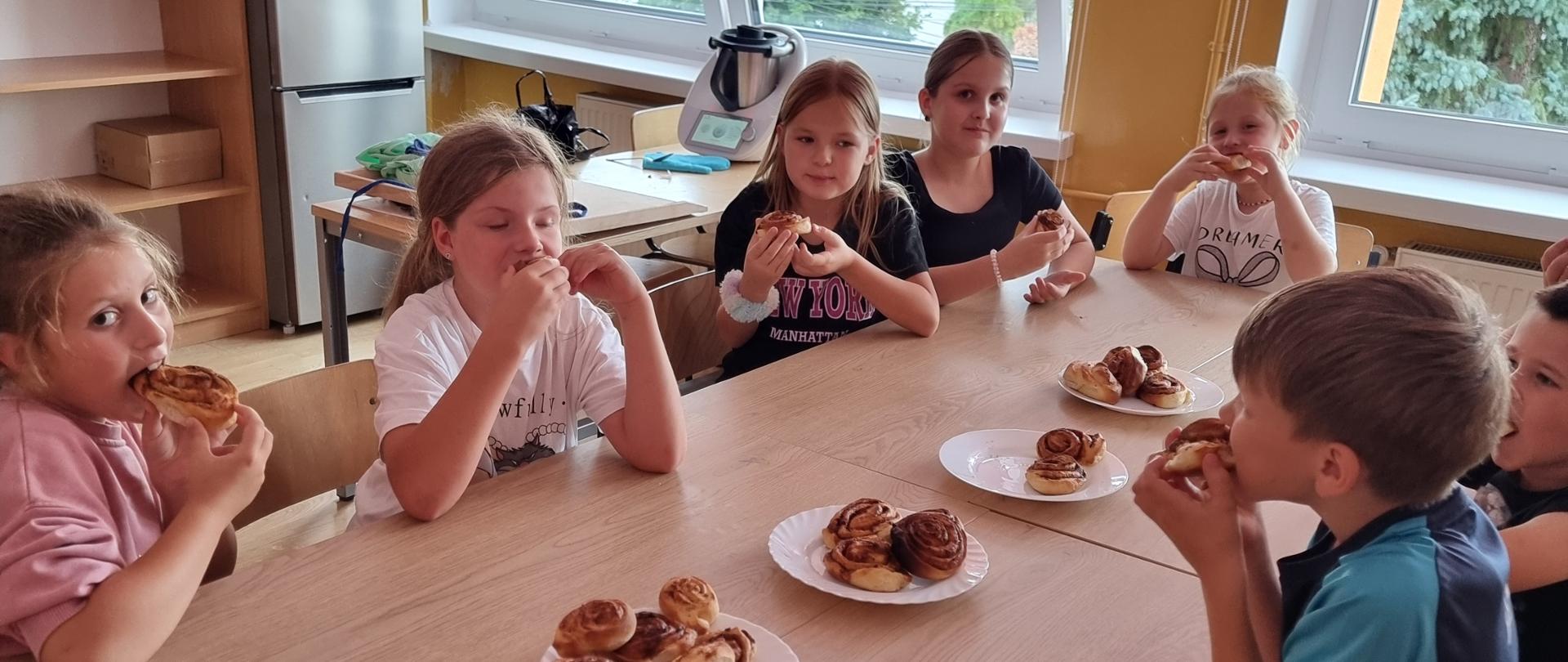 Uczniowie siedzą wokół stolików i degustują upieczone ciasteczka 