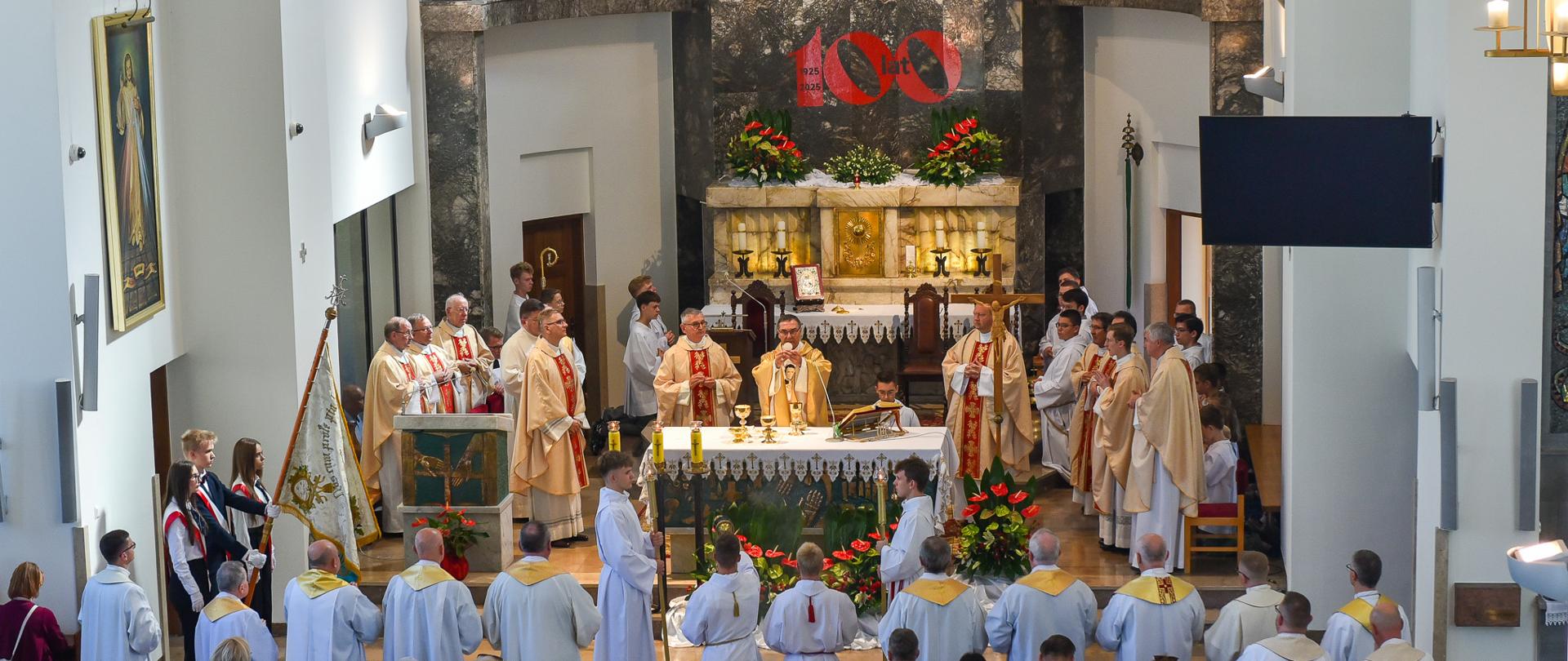 Widok z perspektywy chóru na ołtarz przy którym odbywa się ofiarowanie. Wszyscy klęczą, sprawujący mszę księża w pozycji stojącej patrzą na wznoszącego w górę Ciało Chrystusa Biskupa Piotra Sawczuka.