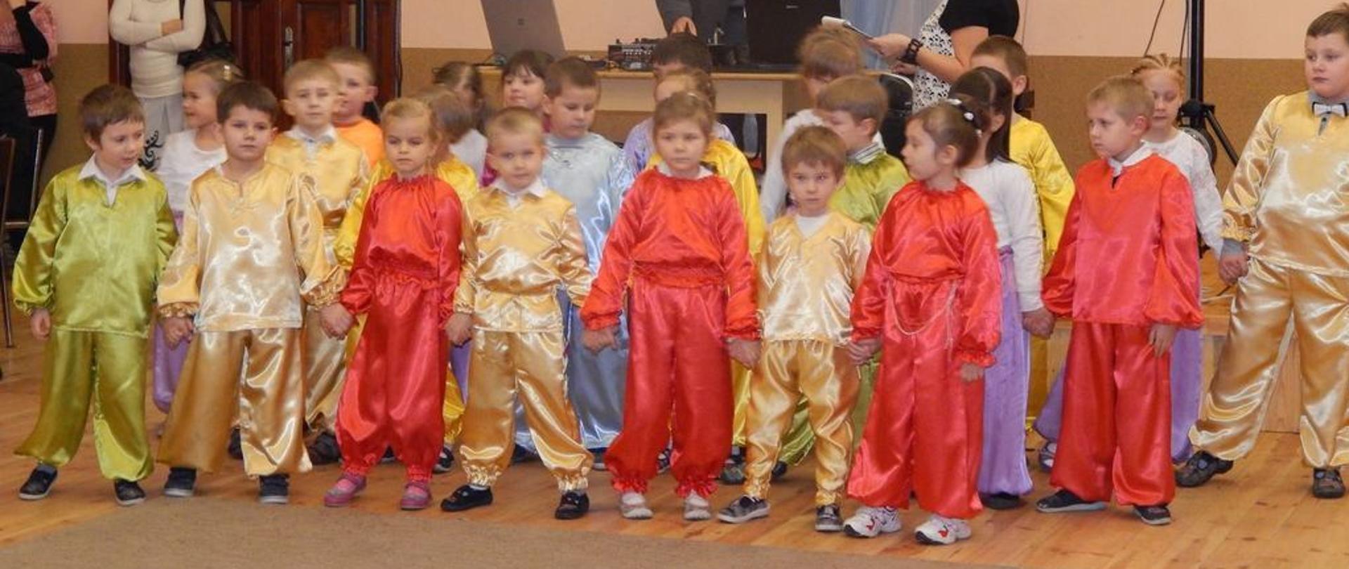 Grupa dzieci w kolorowych świecących strojach stoi w trzech rzędach trzymając się za ręce. Za nimi mężczyzna i kobieta. 