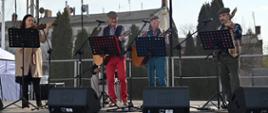 Na zdjęciu widoczny jest zespół muzyczny. Na scenie stoi trzech mężczyzn, którzy grają na gitarach oraz jedna kobieta, grającą na skrzypcach.