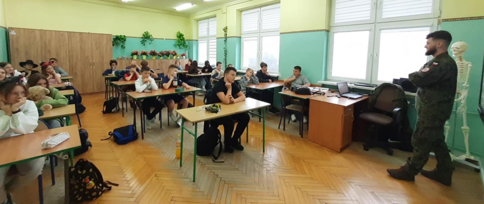 W sali lekcyjnej uczniowie siedzą w ławkach. Przed nimi stoi żołnierz i mówi coś do nich.