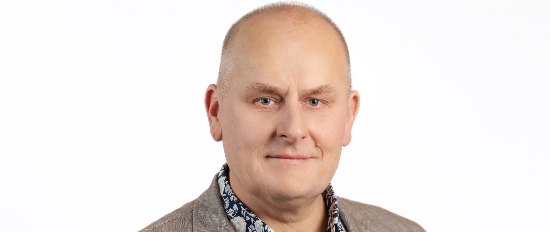 Mężczyzna w średnim wieku lekko łysiejący, łagodna twarz, jasne oczy, lekki uśmiech. Ubrany w jasno brązową marynarkę i granatowa koszulę w białe kwiaty