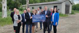 Zdjęcie przedstawia Ministra Macieja Małeckiego oraz Starostę Krzysztofa Bieńkowskiego trzymających wspólnie symboliczną promesę przyznania dofinansowania do budowy Środowiskowego Domu Samopomocy w Przasnyszu. Dookoła nich urzędnicy Starostwa Powiatowego w Przasnyszu oraz podległych mu jednostek.