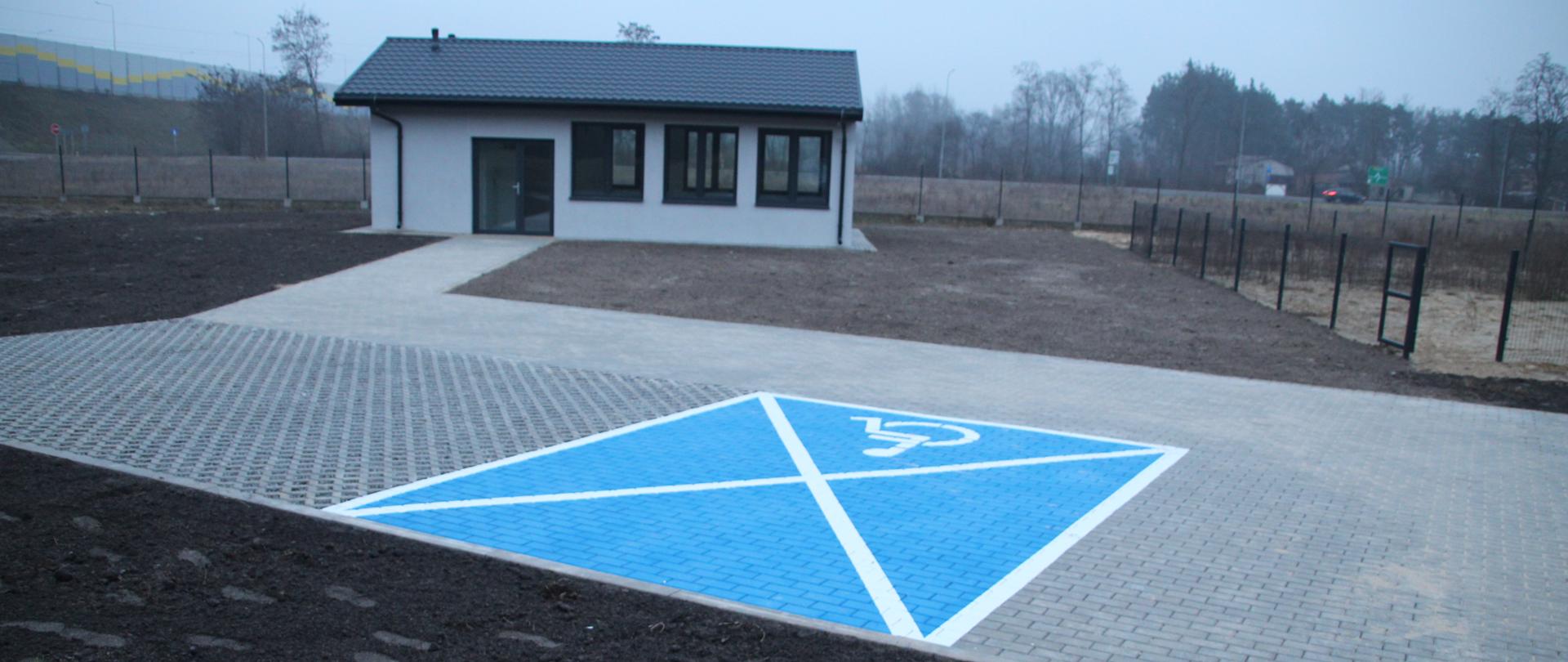 Widok z zewnątrz na nowo wybudowaną świetlicę wiejską w Pawłówku. Jest to parterowy budynek o jasnej elewacji z ciemnym dachem i dużymi oknami. Przed budynkiem znajduje się utwardzony podjazd z kostki brukowej oraz wyraźnie oznakowane, jaskrawoniebieskie miejsce parkingowe dla osób z niepełnosprawnościami z symbolem wózka inwalidzkiego. Teren wokół budynku jest wyrównany, przygotowany pod trawnik.