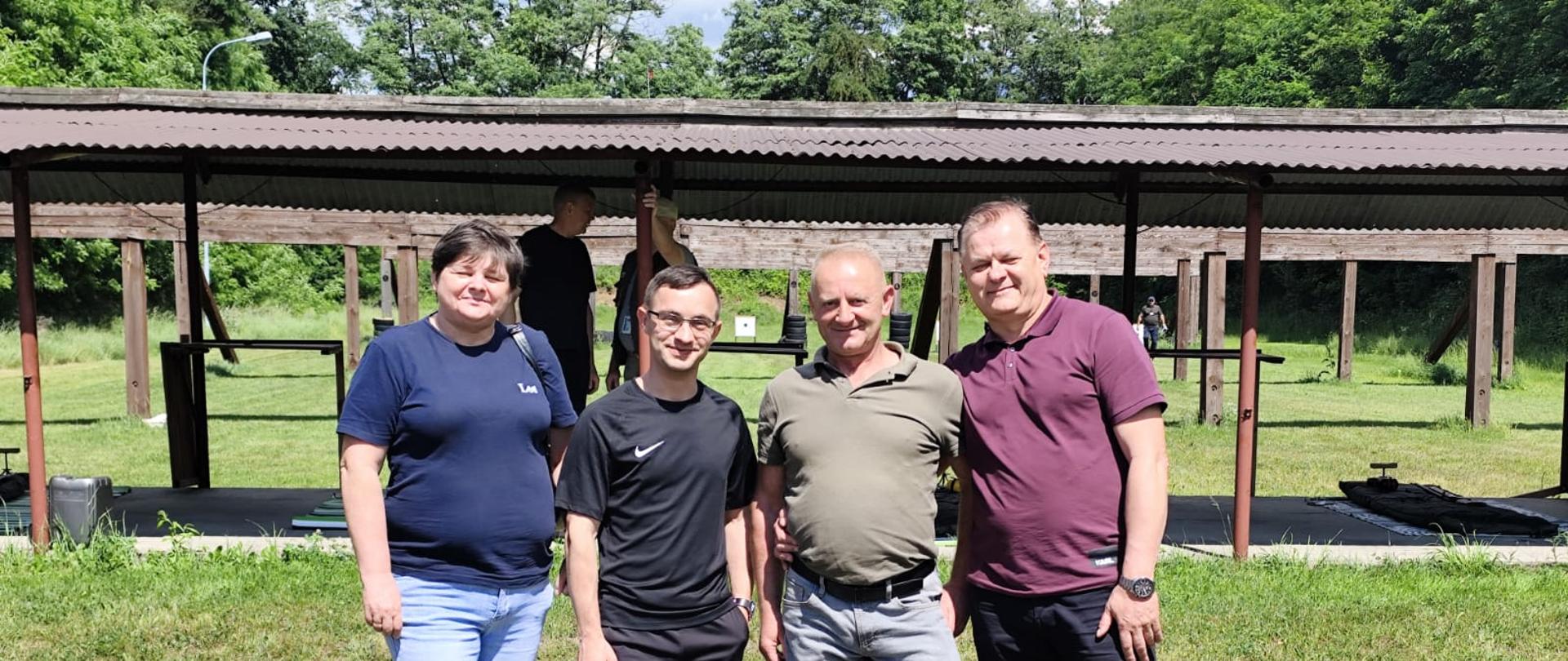 Bliższy kadr przedstawiający cztery uśmiechnięte osoby (kobieta i trzech mężczyzn) stojące na trawie na tle zadaszenia strzelnicy. Pozują zrelaksowani, patrząc bezpośrednio w stronę obiektywu w jasny, słoneczny dzień.