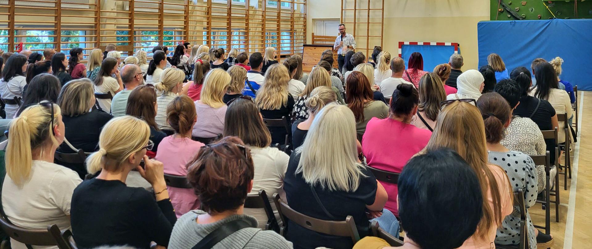 Rodzice siedzący na krzesłach podczas warsztatów profilaktycznych na sali gimnastycznej w szkole