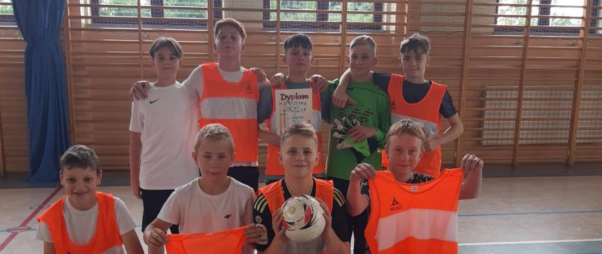 Reprezentacja piłkarska naszej szkoły ustawiona w całym składzie drużyny na hali sportowej w w pomarańczowych koszulkach.