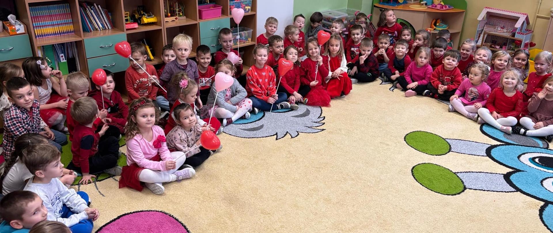 Grupa dzieci w przedszkolu, siedząca w dużym kole na kolorowym dywanie. Większość dzieci ubrana jest na czerwono z okazji Walentynek. Niektóre przedszkolaki trzymają w rękach czerwone i różowe baloniki w kształcie serc.