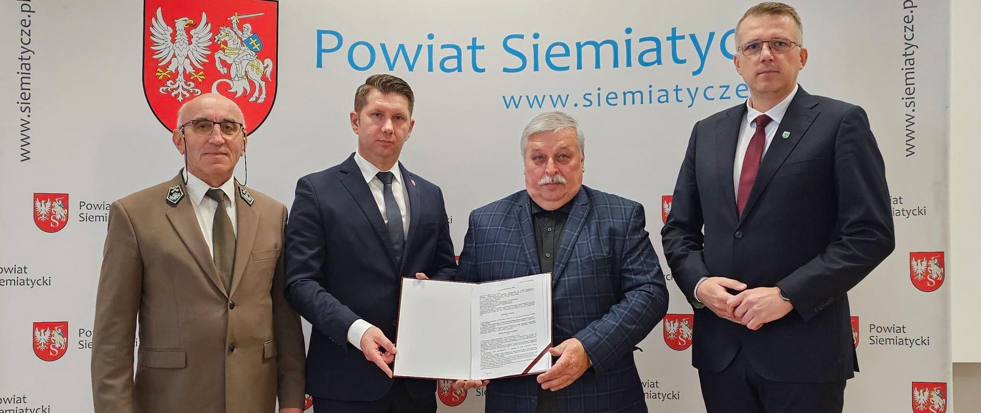 Grupa czterech mężczyzn stoi razem przed transparentem z napisem „Powiat Siemiatycki”. Jeden z mężczyzn trzyma w ręku dokument.
