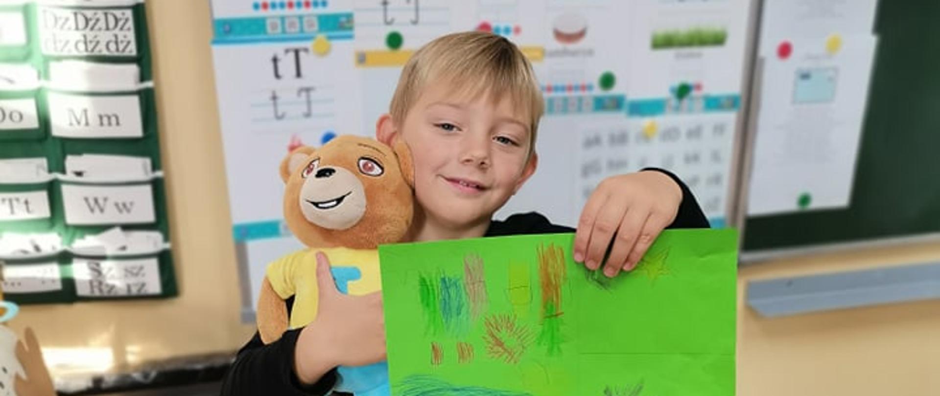 Chłopiec trzyma w prawej ręce pluszowego misia a w lewej zieloną kartkę z rysunkiem