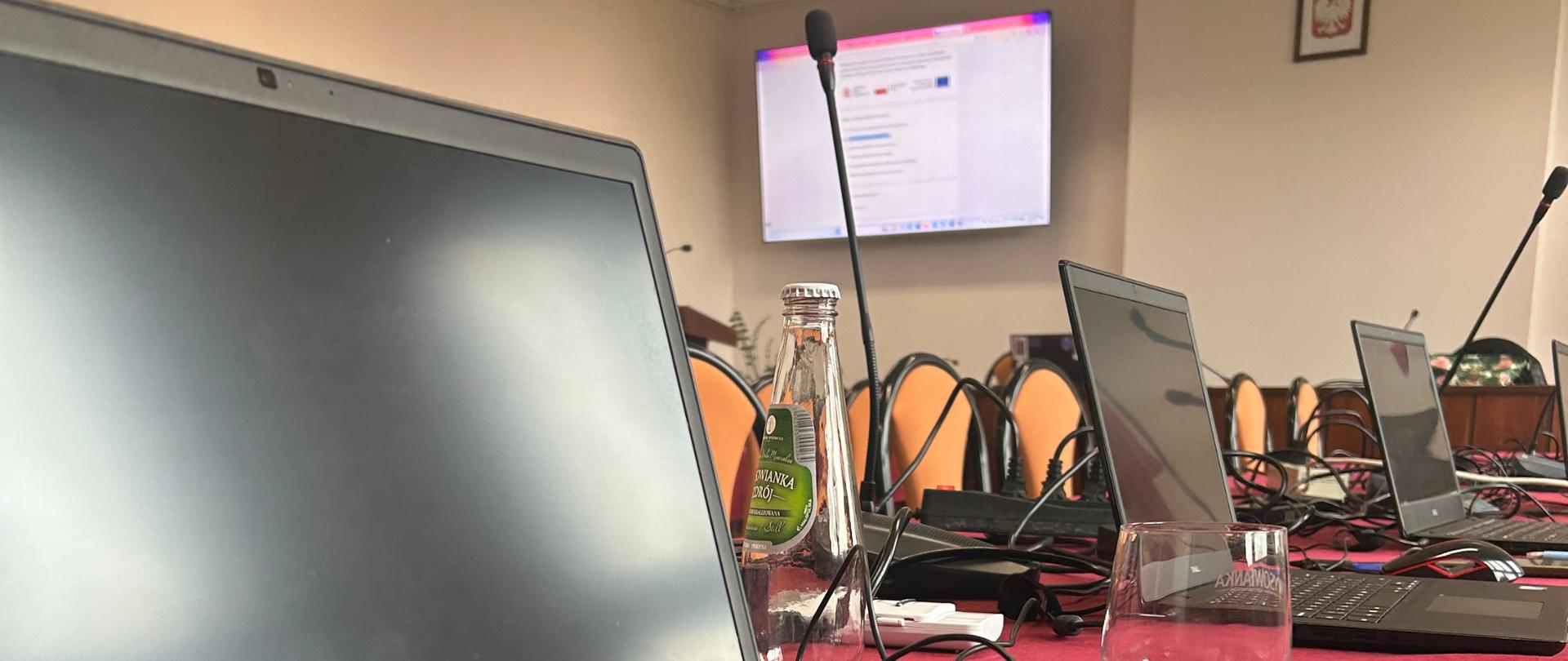 Zbliżenie na laptop Dell stojący na bordowym obrusie w sali konferencyjnej. W tle widać inne laptopy, mikrofony i ekran z wyświetloną stroną internetową.