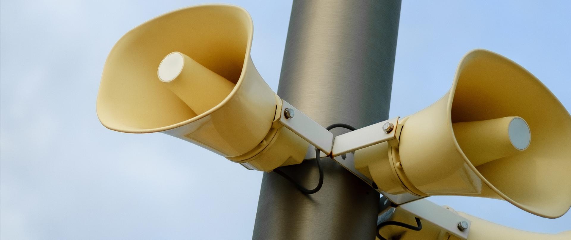 Na tle błękitnego nieba widoczne są żółte syreny alarmowe zamontowane na metalowym słupie. Na grafice umieszczono informację: 5–6 lutego zawyją syreny alarmowe oraz logo Konstantynowa Łódzkiego.
