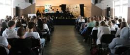 Widok z tyłu auli na scenę, gdzie grupa osób siedzi przy stole. Stół jest przykryty ciemnym obrusem, na którym stoją zapalone lampiony. W tle widać baner z napisem „Gminna Biblioteka w Brańsku” oraz dekorację z cytatem z „Dziadów” Adama Mickiewicza. Aula jest wypełniona rzędami krzeseł, na których siedzi młodzież. Oświetlenie w auli jest naturalne, a na scenie ciepłe i nastrojowe.