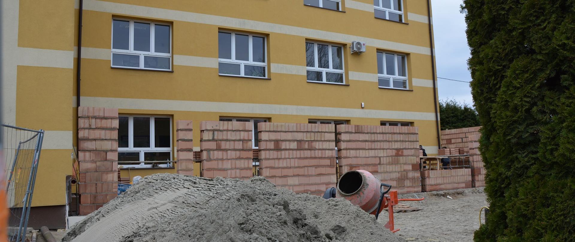 przed żółtym budynkiem szkoły stawiana jest nowa część z czerwonej cegły, przed budynkiem góra pisaku i betoniarka 