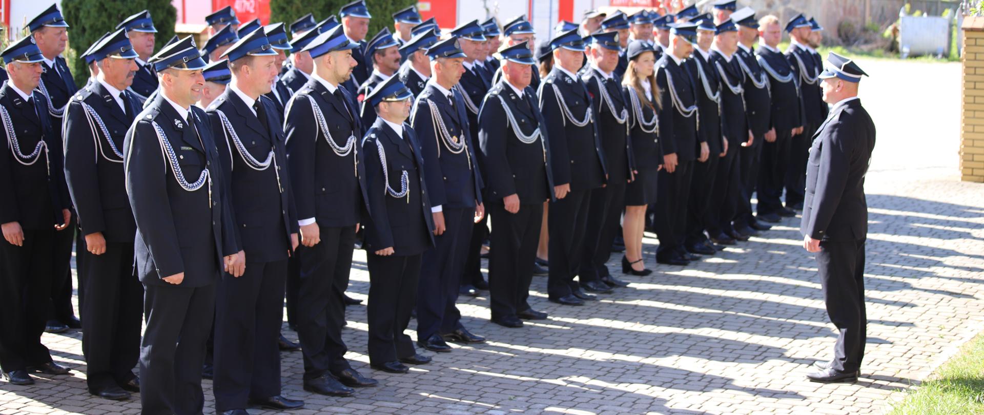 Kobiety i mężczyźni w granatowych mundurach galowych należący do jednostek Ochotniczych Straży Pożarnych z terenu Gminy Brańsk