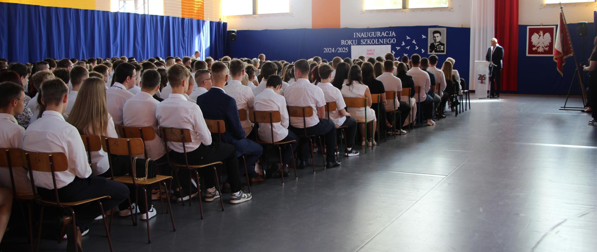 zdjęcie hali podczas rozpoczęcia roku szkolnego , znajdują się uczniowie, a w tle dyrektor ZS1 przemawia