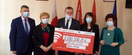 Piątka ludzi w maseczkach. Osoby po środku trzymają duży arkusz z kwotą 1180330,29 zł. Za nimi stoją flagi polski, województwa Mazowieckiego, gminy Zabrodzie oraz flaga Unii Europejskiej