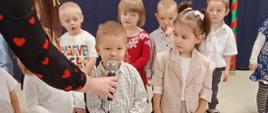 Przedszkolaki stoją w sali na tle ścianki "Pasowanie na przedszkolaka". Przy mikrofonie stoi jeden z przyszłych przedszkolaków.
