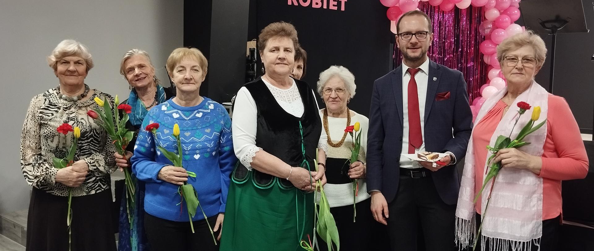 Panie z klubu senior+ pozujące do zdjęcia z burmistrzem Dąbrowy Białostockiej, trzymające goździka i tulipana