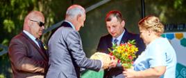 Kadr przedstawiający gest przekazania i dzielenia chleba dożynkowego oraz bukietu polnych kwiatów pomiędzy gospodarzami wydarzenia na scenie.