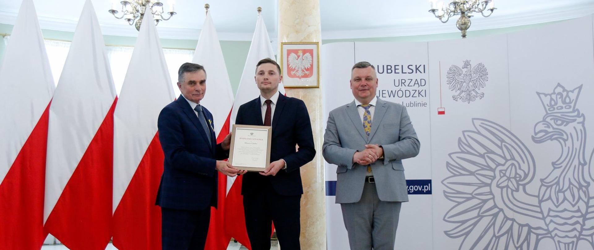 Zdjęcie przedstawia trzy osoby stojące wewnątrz budynku na tle flagi Polski. Jedna z nich trzyma w ręce oprawiony w ramkę dyplom.