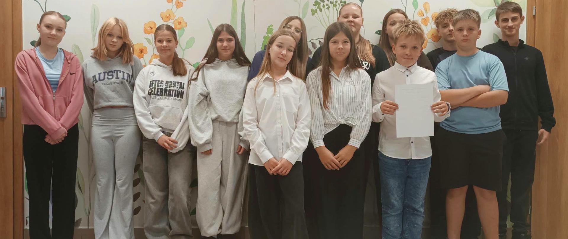 Na zdjęciu widać grupę kilkunastu młodych osób ustawionych wspólnie do fotografii w jasnym, ozdobionym roślinnymi i kwiatowymi motywami korytarzu. Większość osób stoi w dwóch rzędach, a kilka osób znajduje się z przodu.
Uczestnicy mają na sobie głównie codzienne, wygodne ubrania — bluzy, koszule, spodnie dresowe i jeansy. Jedna z osób z przodu trzyma białą kartkę. W tle znajdują się drewniane drzwi oraz duża ścienna dekoracja przedstawiająca kwiaty, liście i motyle.