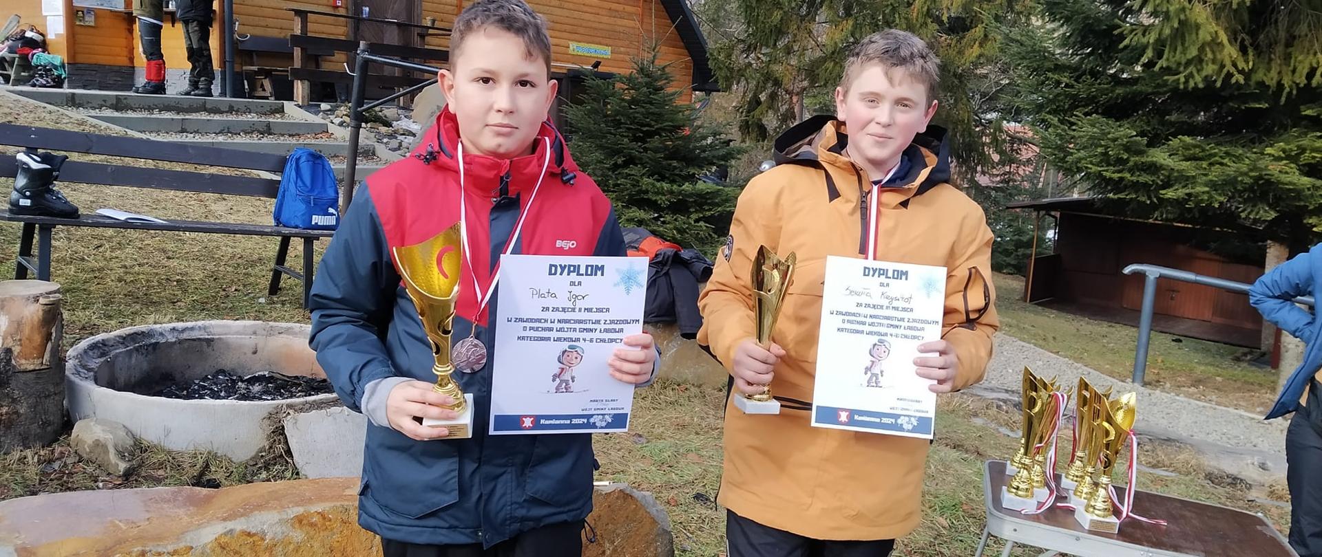 Dwóch uczniów naszej szkoły pozuje w strojach narciarskich z medalami, pucharami i dyplomami zdobytymi w Zawodach narciarskich o Puchar Wójta Gminy Łabowa