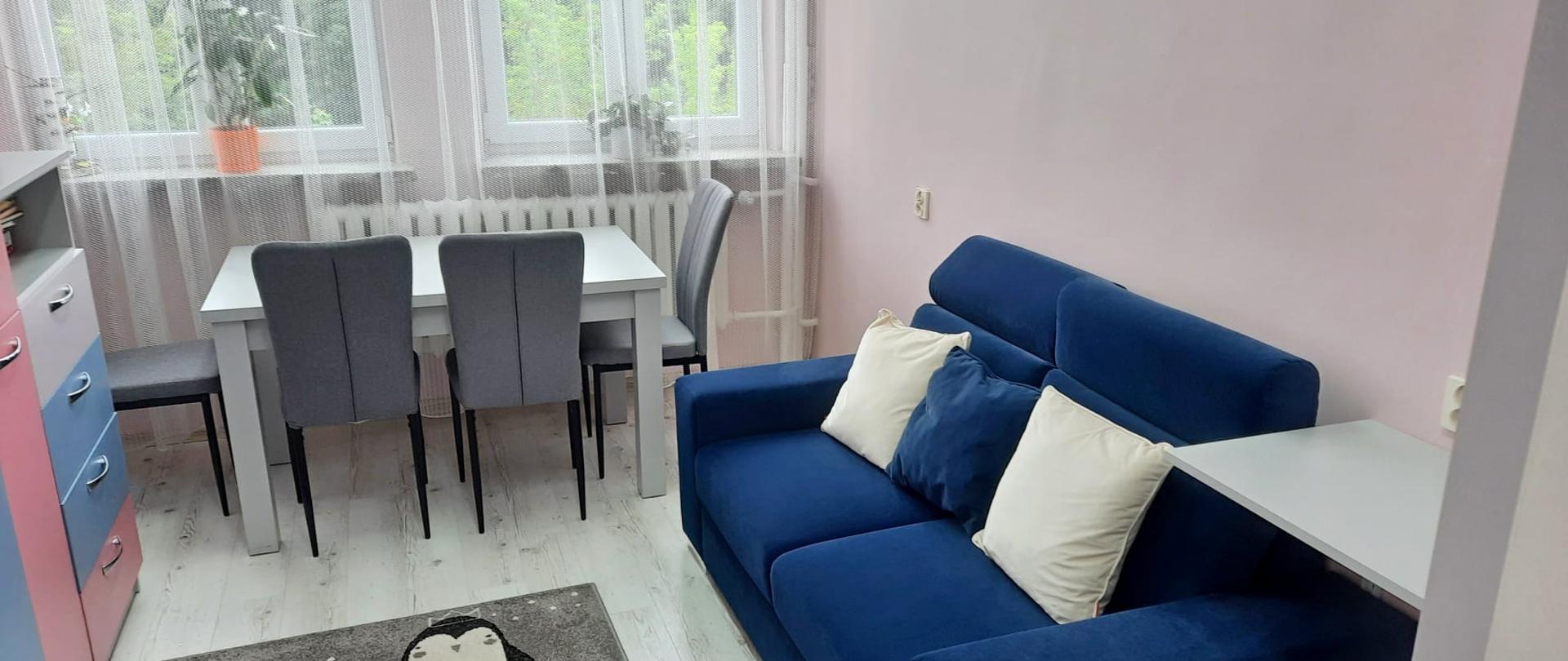 Fotografia przedstawia nowo utworzony w Powiatowym Centrum Pomocy Rodzinie w Przasnyszu pokój do rodzinnych spotkań. Ściany w kolorze delikatnie brzoskwiniowym, po lewej szafki w kolorach jasno szarym, niebieskim i różowym. Po lewej ciemno niebieska wygodna kanapa z dwoma poduszkami w kolorze kanapy oraz jedną białą. Na wprost jasno szary stół obstawiony czterema krzesłami koloru szarego oraz dwa okna z białymi firanami. Na podłodze miękki szary dywan, na którym przedstawiono cztery rysunkowe biało-czarne pingwiny. 