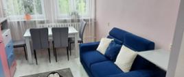 Fotografia przedstawia nowo utworzony w Powiatowym Centrum Pomocy Rodzinie w Przasnyszu pokój do rodzinnych spotkań. Ściany w kolorze delikatnie brzoskwiniowym, po lewej szafki w kolorach jasno szarym, niebieskim i różowym. Po lewej ciemno niebieska wygodna kanapa z dwoma poduszkami w kolorze kanapy oraz jedną białą. Na wprost jasno szary stół obstawiony czterema krzesłami koloru szarego oraz dwa okna z białymi firanami. Na podłodze miękki szary dywan, na którym przedstawiono cztery rysunkowe biało-czarne pingwiny. 