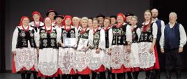 kobiety i mężczyźni w pięknych ludowych strojach w kolorach białych czarnych i czerwonych stoją na scenie i pozują do wspólnego zdjęcia 