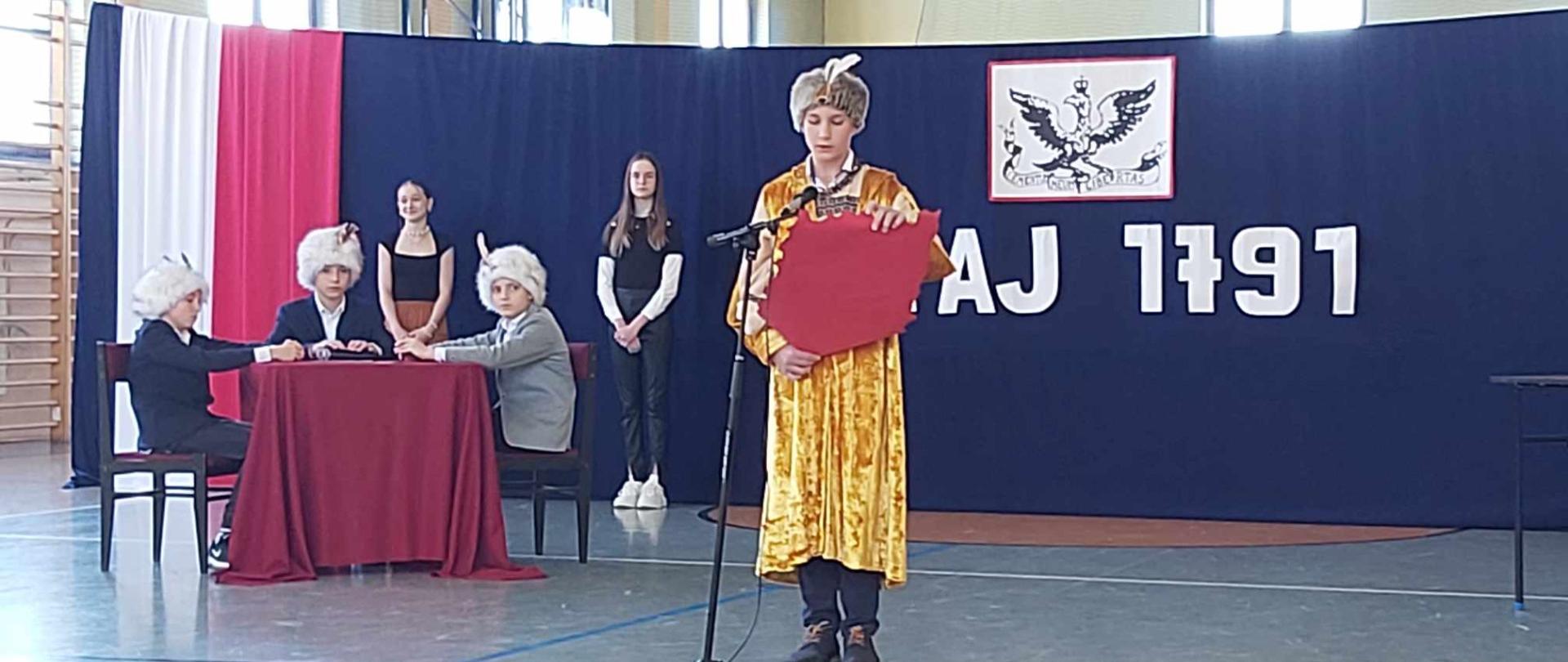 Uczniowie w przepięknych strojach na tle dekoracji 3 majowej z napisem w tle "Maj 1791'"