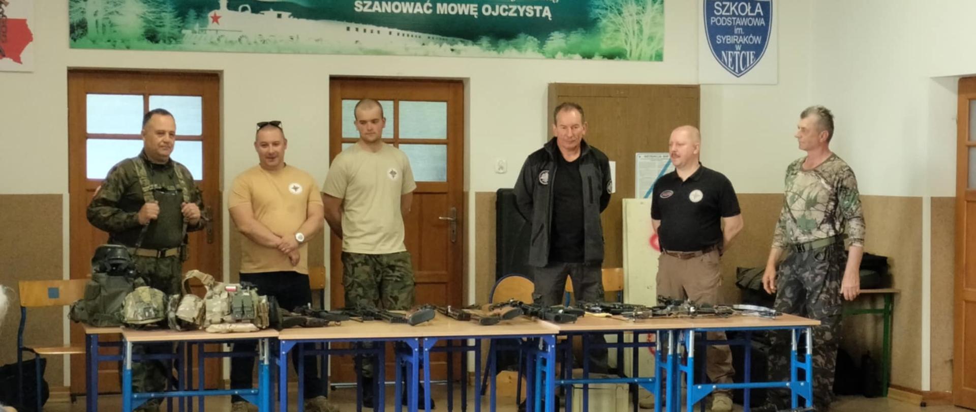 Sześcioro mężczyzn stoi przed stołami na których leżą różne typy broni palnej.