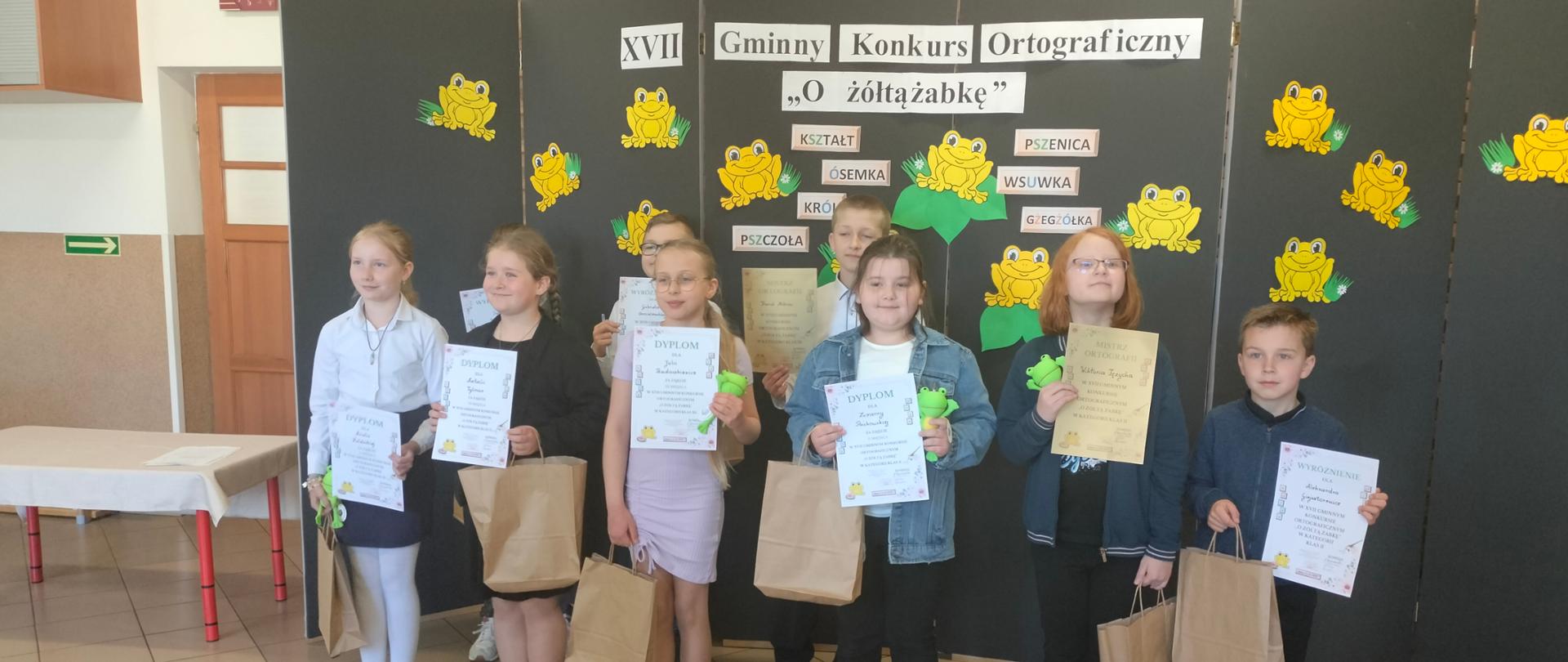 Dziewięcioro uczniów pozuje do wspólnego zdjęcia, W dłoniach trzymają dyplomy oraz torebki prezentowe. Za nimi stoi dekoracja na której wisi napis: ,,XVII Gminny Konkurs Ortograficzny ,,O żółtą żabkę".