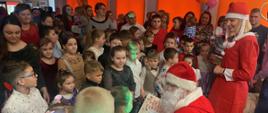 Na zdjęciu znajduje się Mikołaj, który siedzi na krześle, a w ręku trzyma prezent. Obok niego stoi kobieta ubrana za w czerwoną sukienkę i mikołajkową czapkę. Wokół nich stoją dzieci oraz rodzice.