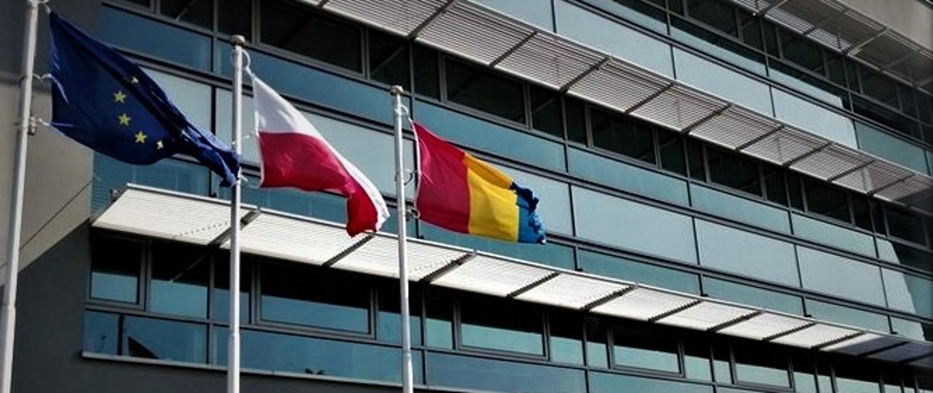 Szary budynek z oknami , na pierwszym planie trzy maszty z flagami , od lewej flaga Unii Europejskiej, w środku flaga Polski i flaga powiatu polkowickiego. 