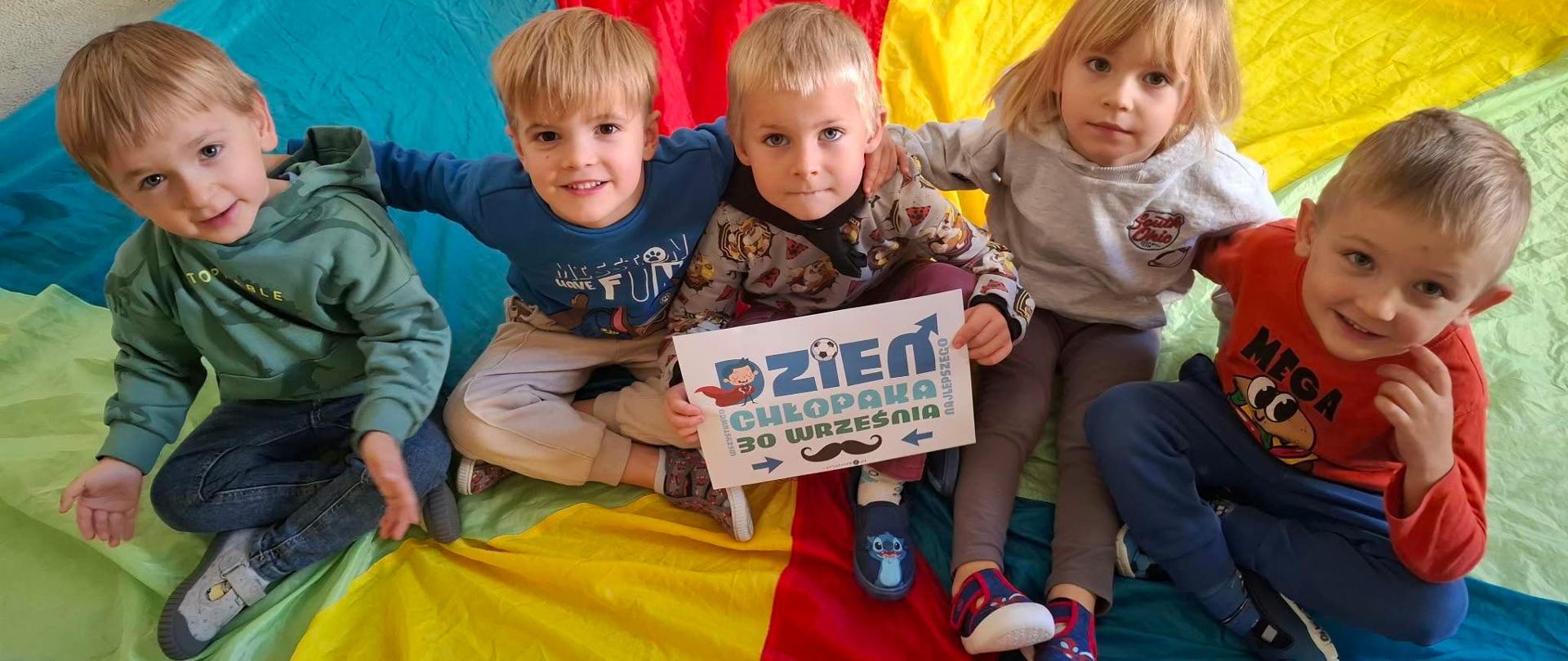 Na zdjęciu widać grupę pięciorga małych dzieci siedzących razem na dużej, kolorowej chuście. Dzieci uśmiechają się lub patrzą w stronę aparatu i mają na sobie wygodne, codzienne ubrania w różnych kolorach.
W centrum jedno z dzieci trzyma kartkę z napisem „DZIEŃ CHŁOPAKA – 30 WRZEŚNIA”, co wskazuje, że fotografia została wykonana z okazji tego święta. W tle również widać kolorowe fragmenty materiału oraz częściowo kolejne dziecko. Całość ma radosny, przedszkolny charakter.
