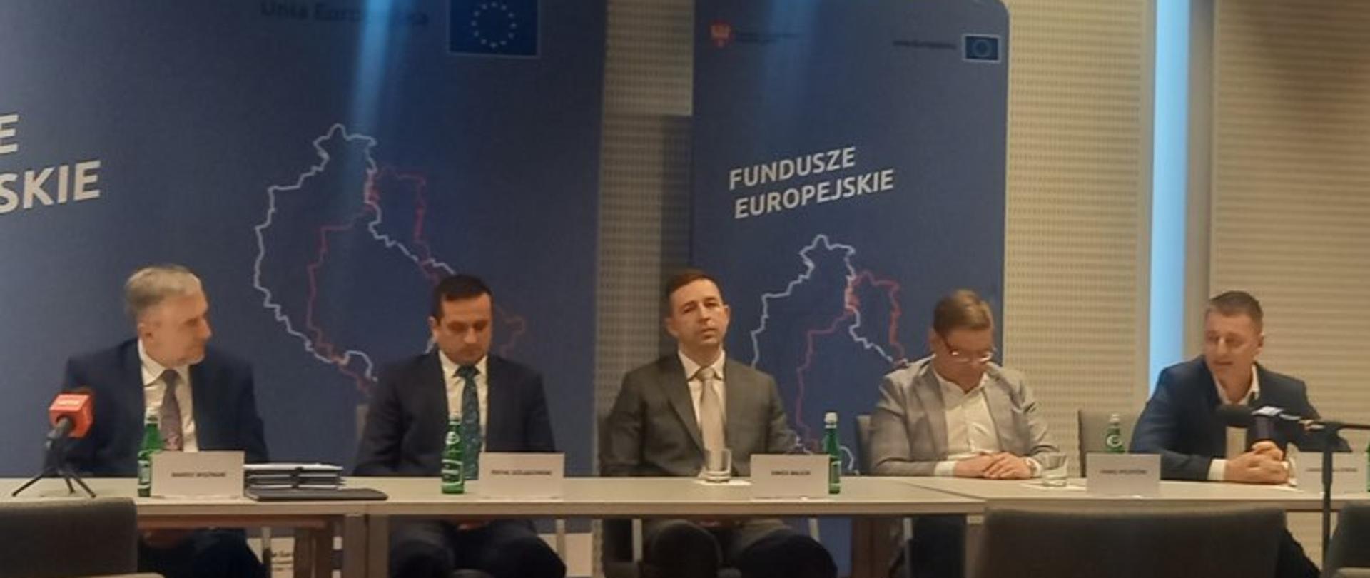 5 osób siedzących za stołem w sali konferencyjnej, w tle banery z logotypami i napisami Fundusze Europejskie